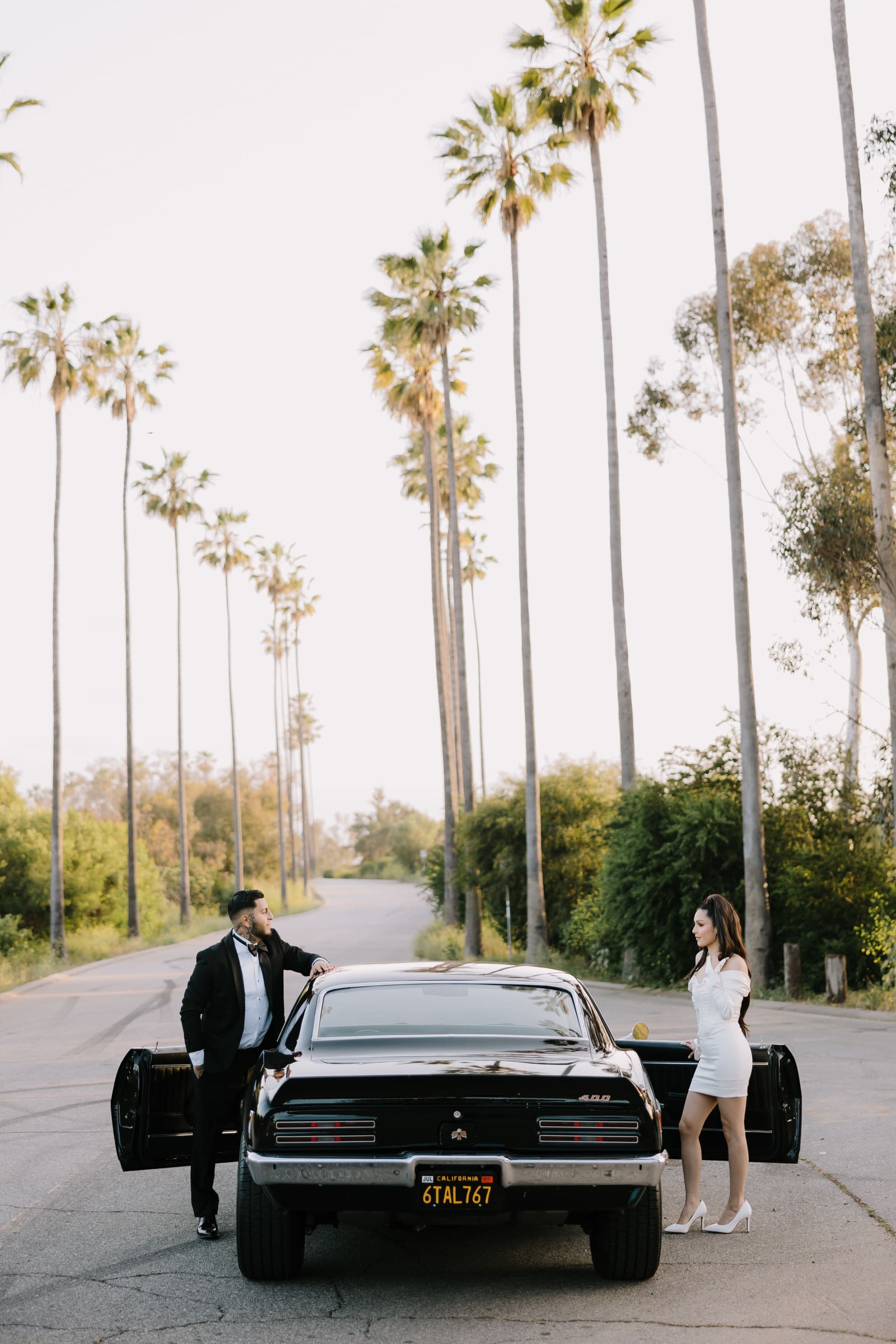 Alejandra and Jovanny leaning against a black classic Firebird with the DTLA skyline and palm trees at Elysian Park.