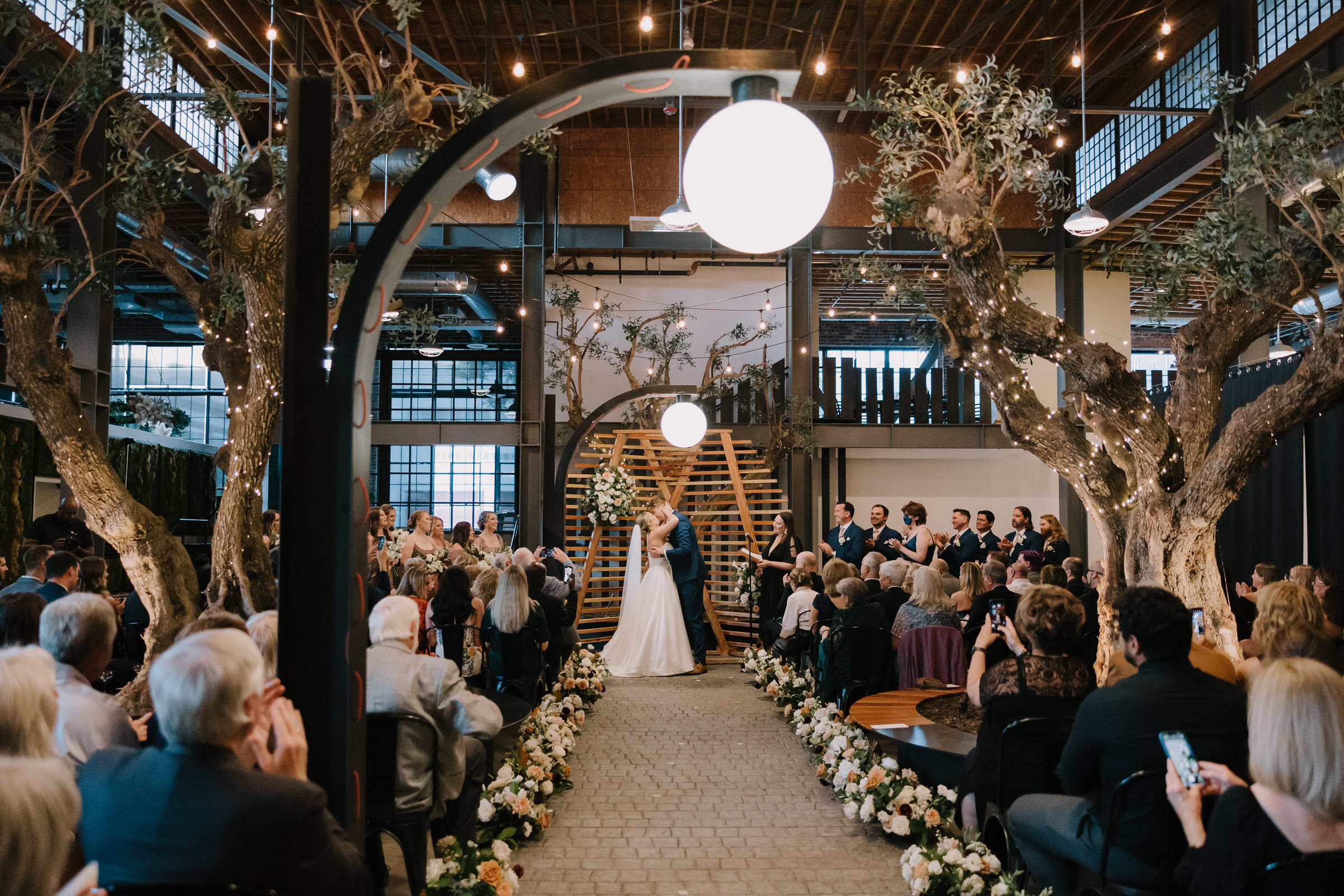 A joyful shot of Melissa and Ari during their wedding ceremony under the fairy-lit olive trees at Venue 808 in San Dieg