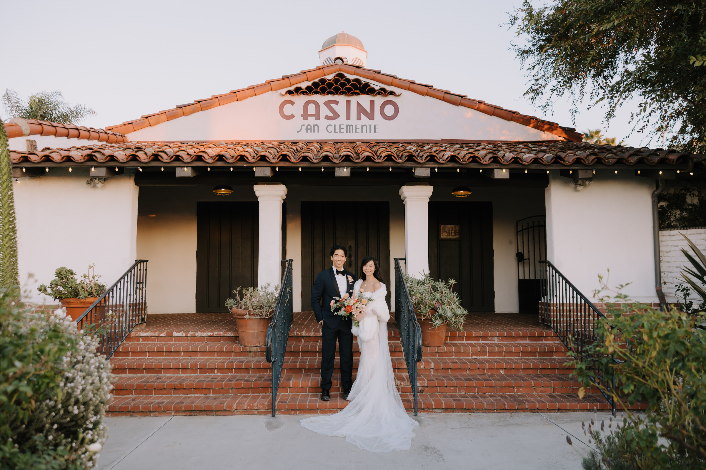 Angela and Brandon standing in front of the historic white-walled architecture of the Casino San Clemente during their wedding.